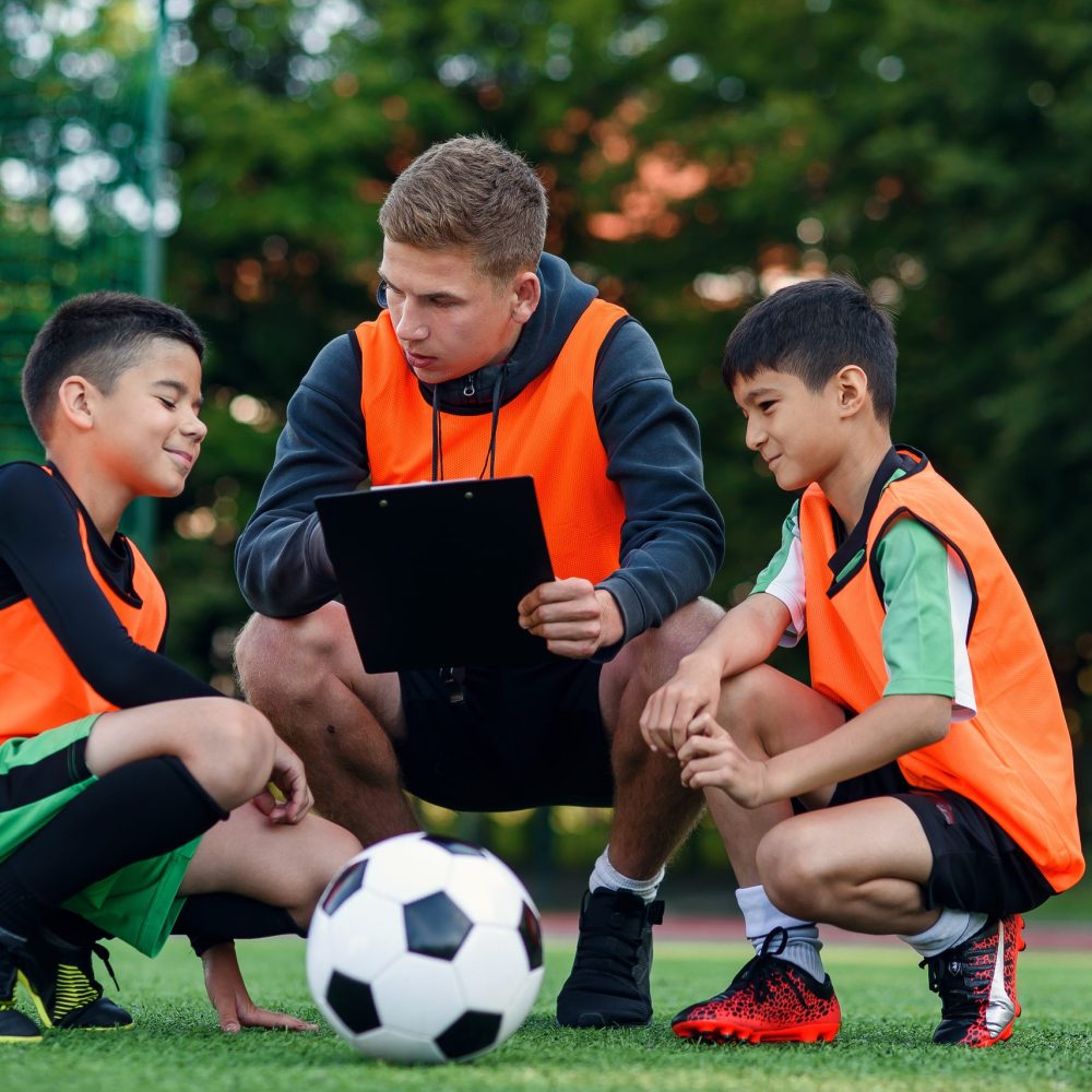 Professional young soccer coach tells the strategy of football game to his attentive teen players at stadium during training.