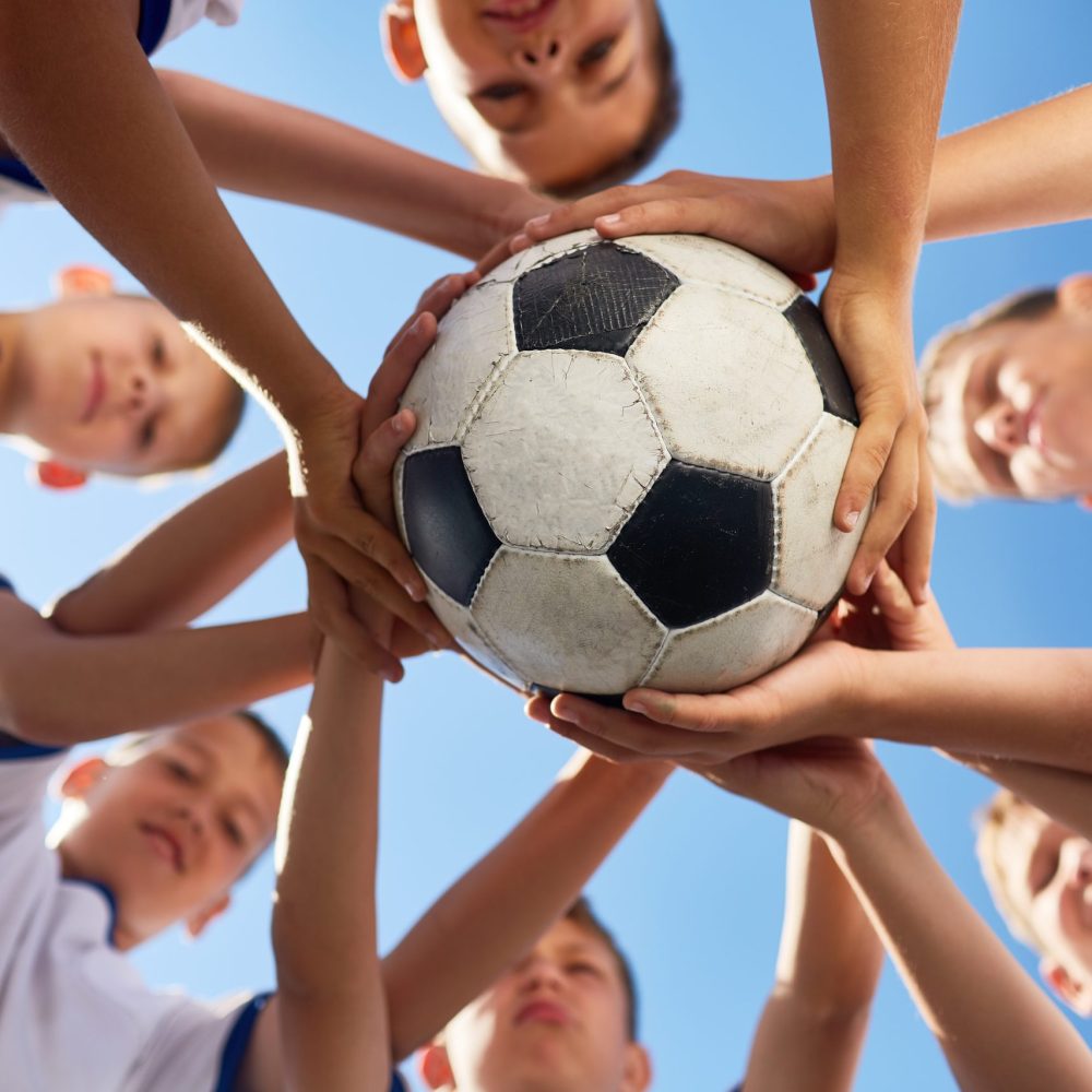 Low angle view of boys in junior football team standing in circle holding ball together against  blue sky, focus on ball